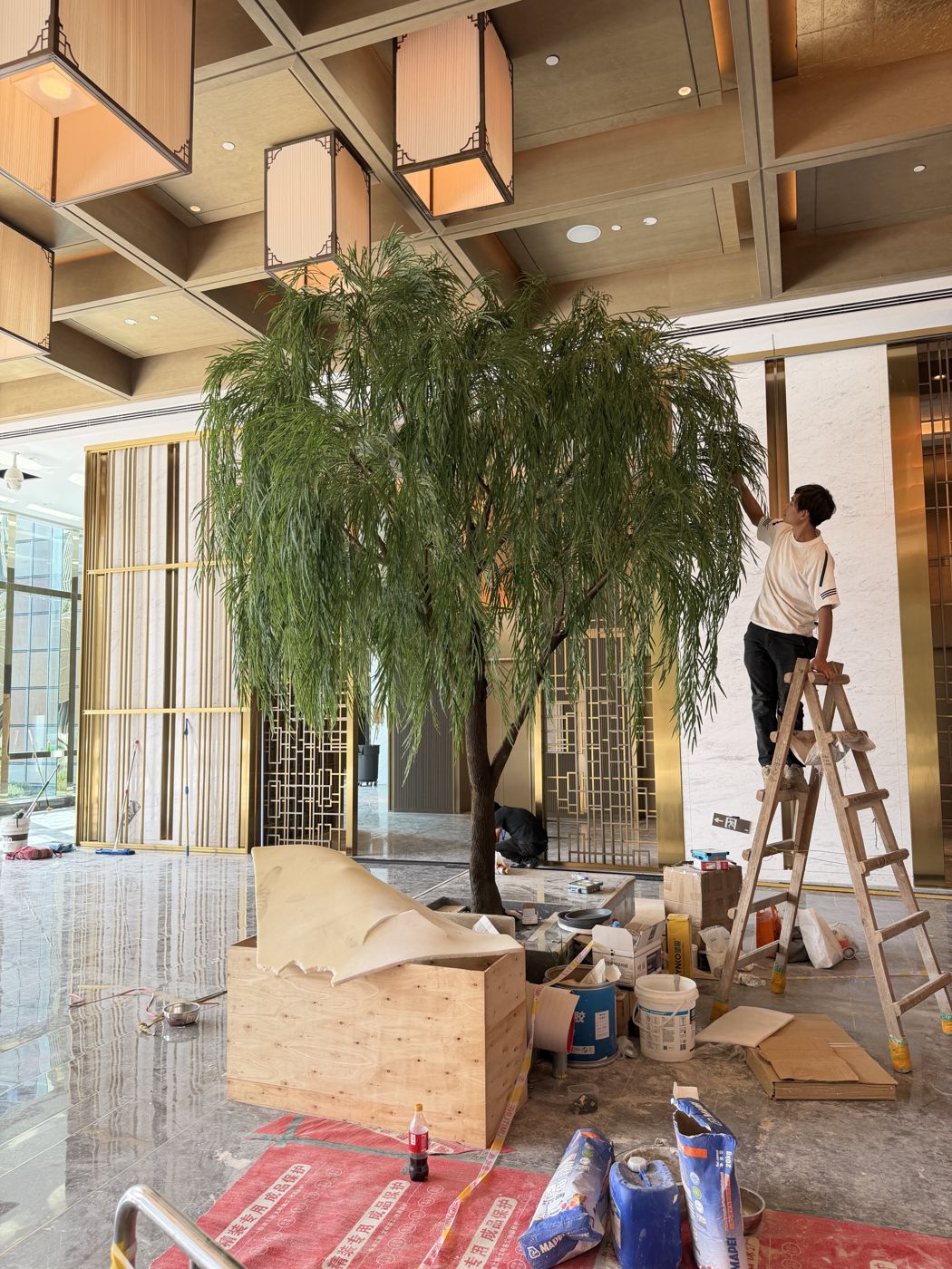 An installer on a ladder shaping an artificial willow in a hotel lobby